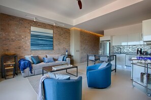 A modern living room with a sofa, coffee table, and blue armchairs. A kitchen with a refrigerator and coffee maker is visible in the background. - Rooftop Pool, Gym & Hot Tub | Stylish 1BR Loft (Puerto Vallarta)