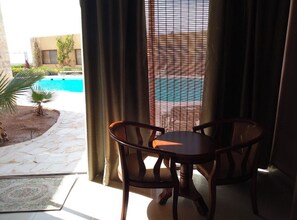 A room with a view of a swimming pool, a table with two chairs, and a window with blinds. - Bungalow in Aqaba Gulf Red Sea (Aqaba)