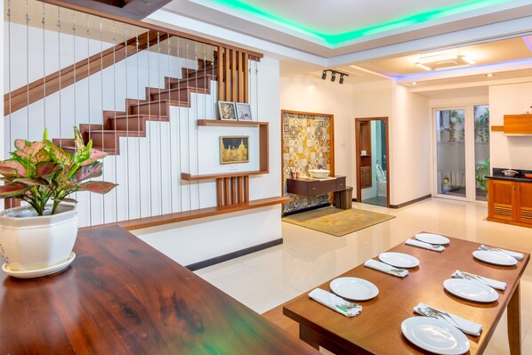 A modern dining area with a wooden table set for six, a staircase with wooden railings, and a kitchen visible in the background.