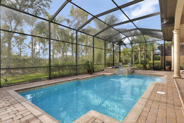 Uma piscina interior com estrutura de vidro, rodeada por um pátio de tijolos e plantas em vasos.