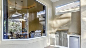 An outdoor kitchen with a grill, refrigerator, and dining area visible through a large window. - A Rare Find! Lely Resort Single Family Home in Prestigious Majors Community (Naples)