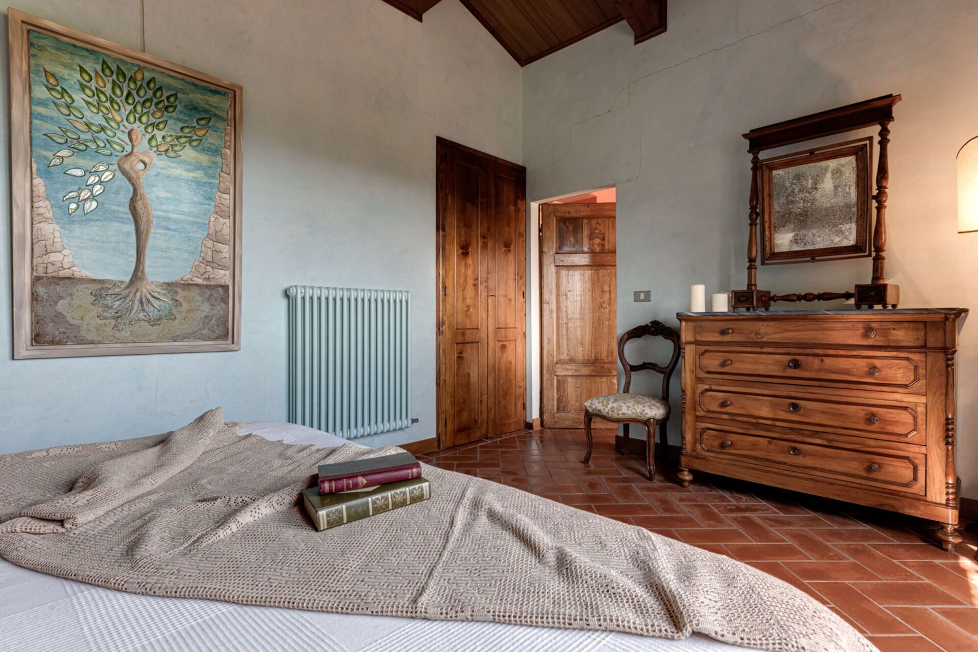 Een slaapkamer met een bed, een dressoir, een stoel, een radiator en een schilderij van een boom.