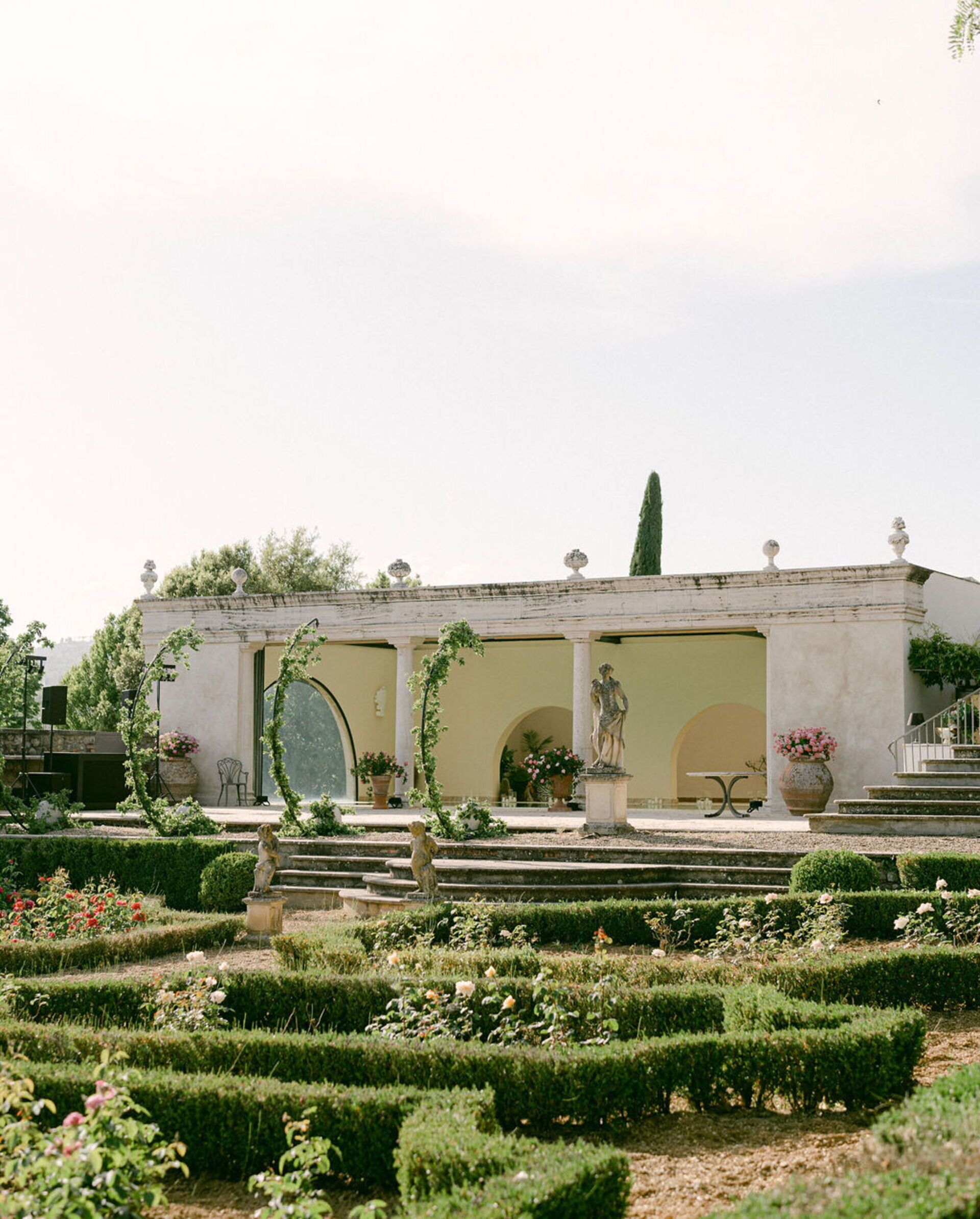 A classical building with a garden featuring a statue, a fountain, and neatly trimmed hedges.