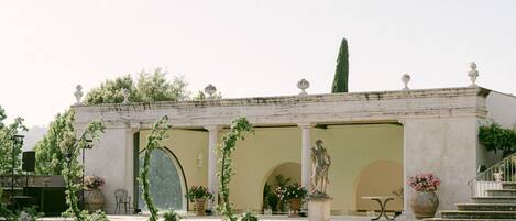 A classical building with a garden featuring a statue, a fountain, and neatly trimmed hedges.