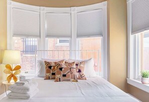 Una camera d'albergo con un letto, cuscini, una lampada e una finestra con persiane.