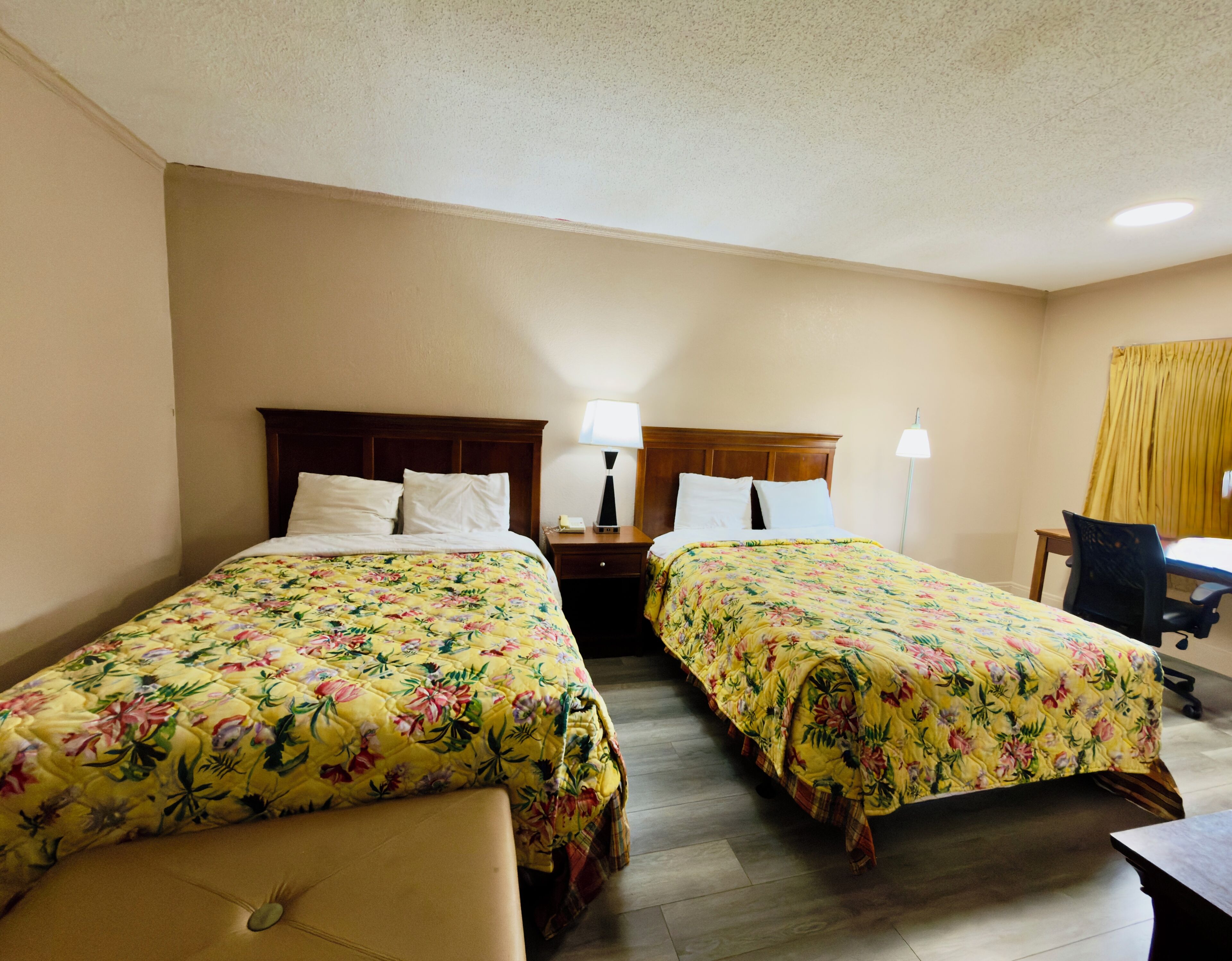 A hotel room with two beds, each with floral bedding, a nightstand with a lamp, a desk with a chair, and a window with yellow curtains.
