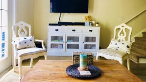 A living room with a television mounted on the wall, a white cabinet with multiple drawers, two white chairs with patterned cushions, and a wooden coffee table with a candle.