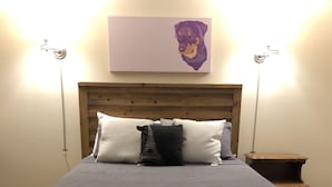 A bedroom with a wooden bed, grey bedding, a bench, and a wall-mounted lamp.