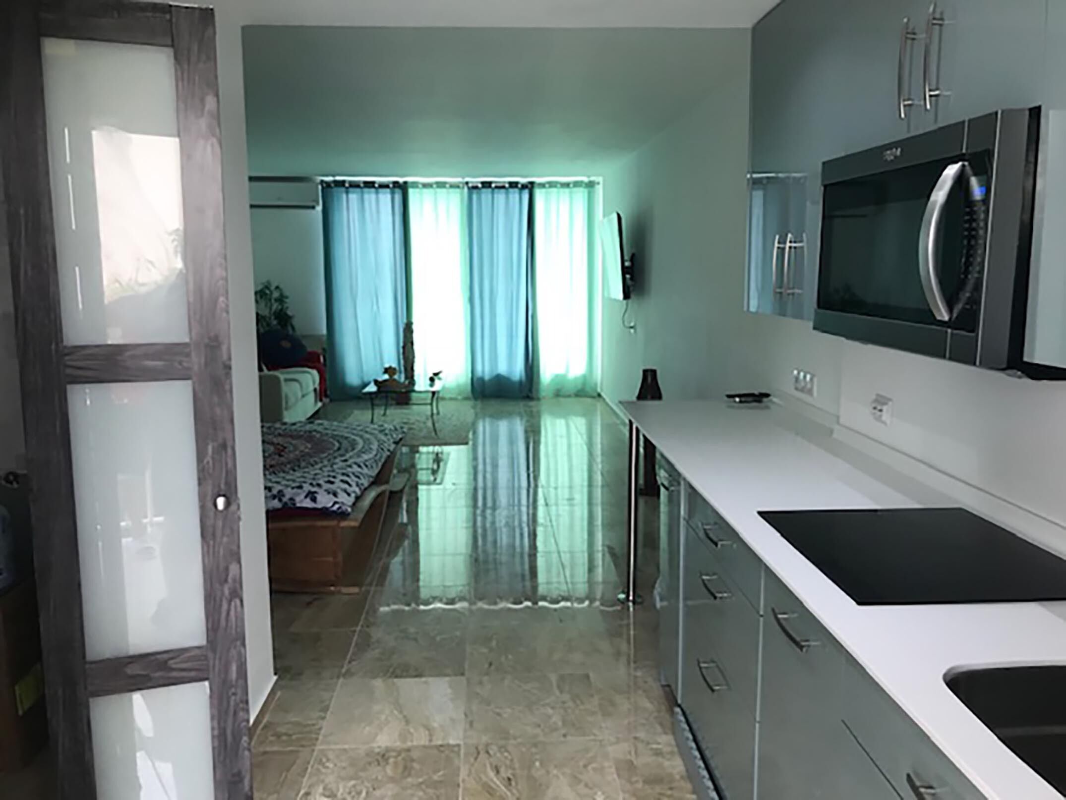 A modern kitchen with a large refrigerator, a microwave, and a sink. There is a dining area with a table and chairs. The room has a large window with curtains.