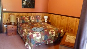 A bedroom with a wooden headboard, a quilted bedspread, two rolled towels, a bedside lamp, and a nightstand with a lamp. - Spacious fully self contained two bedroom chalet, nestled in the bush. (Deepdene)