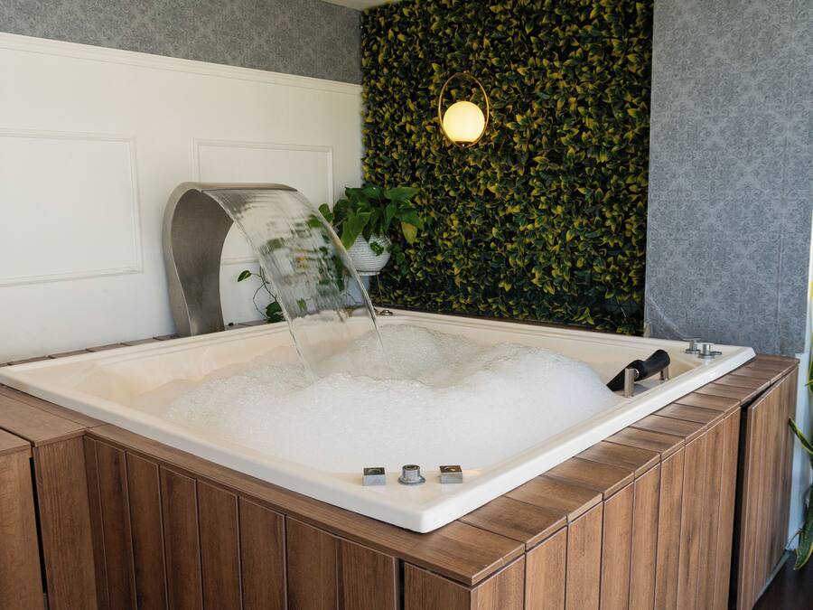 A modern bathroom with a wooden bathtub, a wall-mounted light fixture, and a green leafy wall.