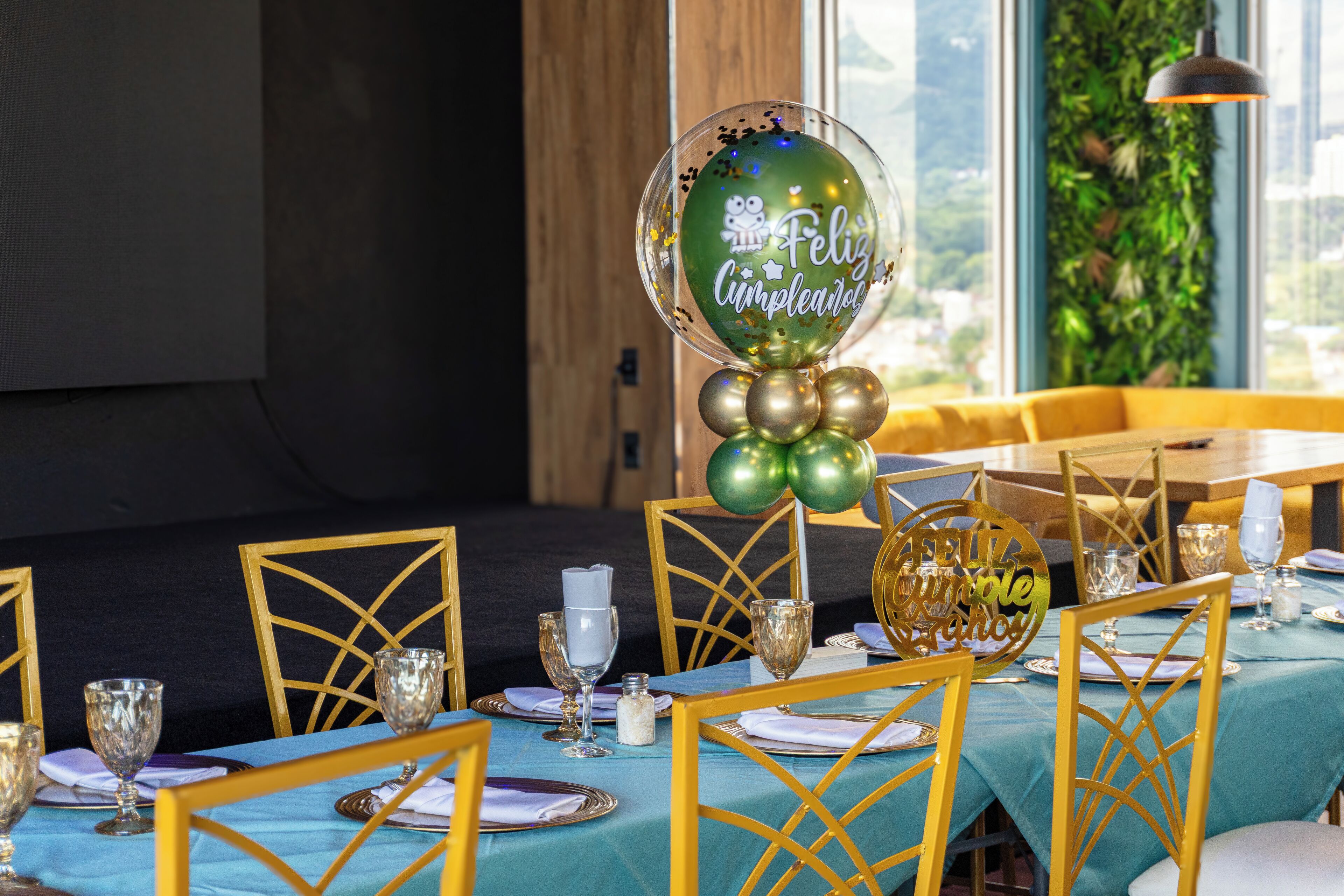 Un comedor con mesas dispuestas para una celebración, con sillas doradas, manteles azules y un globo verde grande con el texto "Feliz Cumpleaños".