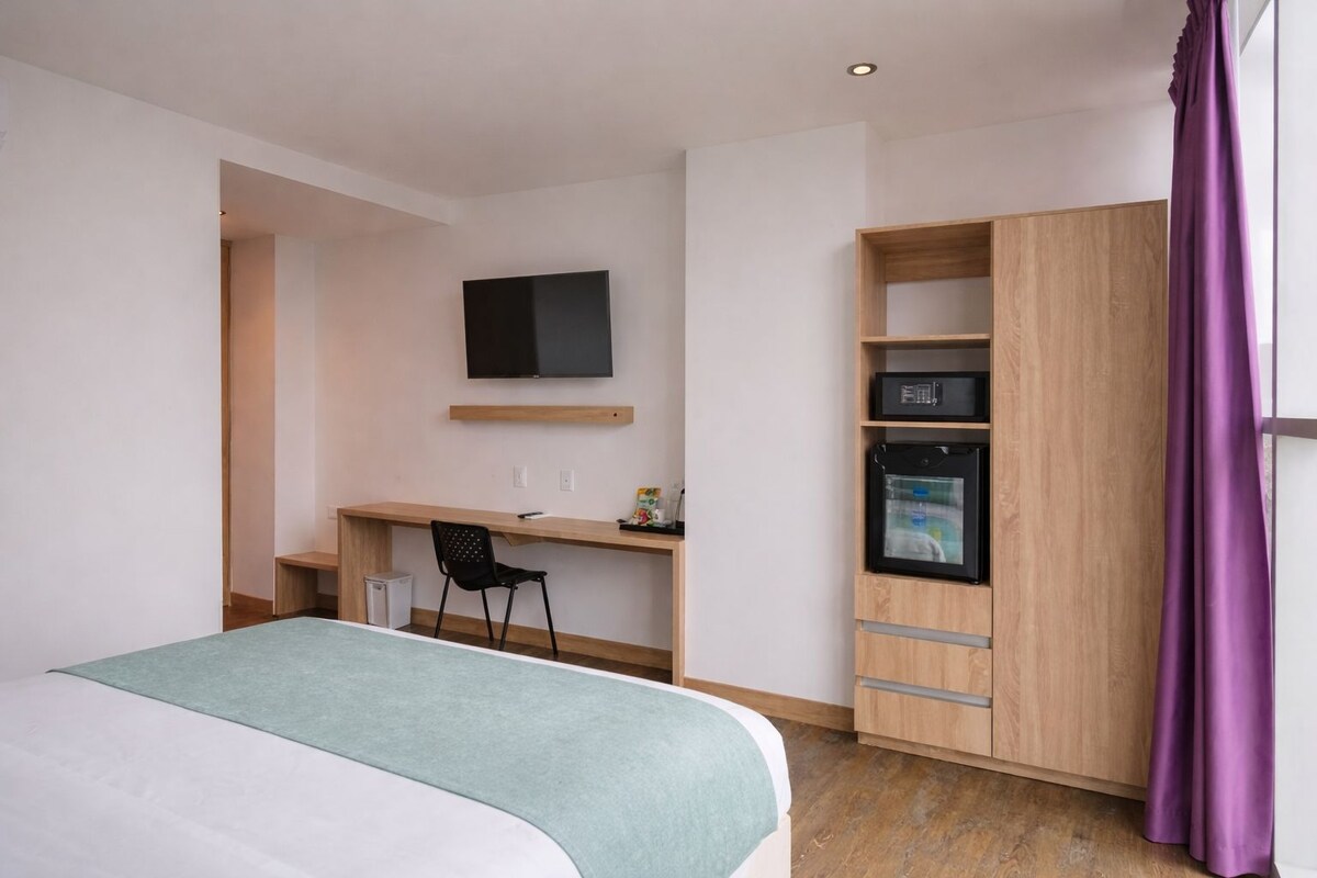 A hotel room with a bed, a desk with a chair, a television, a minibar, and a wardrobe.