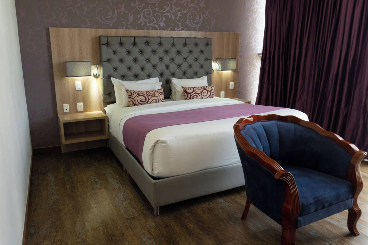 A neatly arranged hotel room with a large bed, two bedside tables with lamps, a wooden headboard, and a classic armchair.