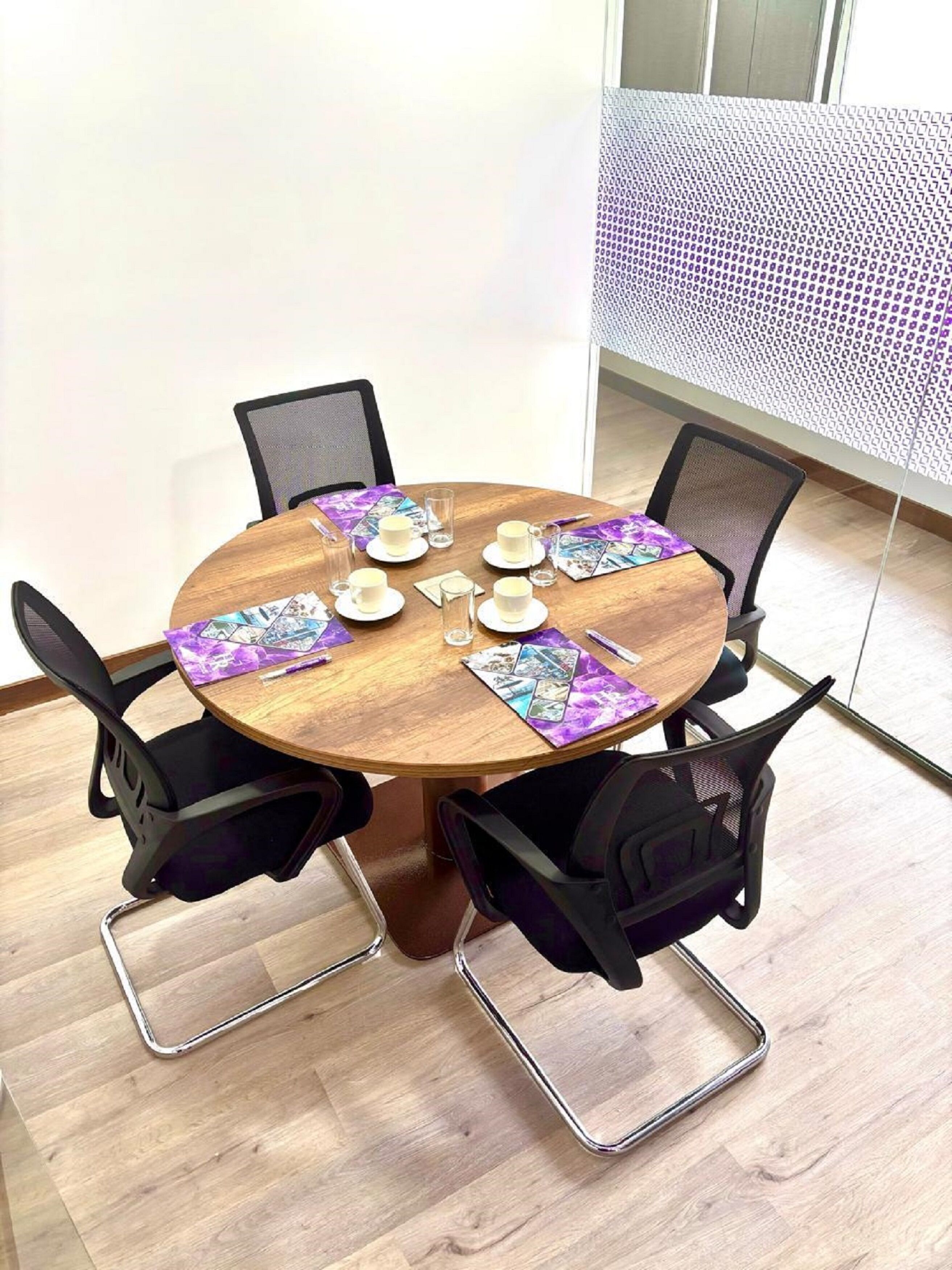 A round wooden table with four black chairs, a purple placemat, and a white tablecloth.