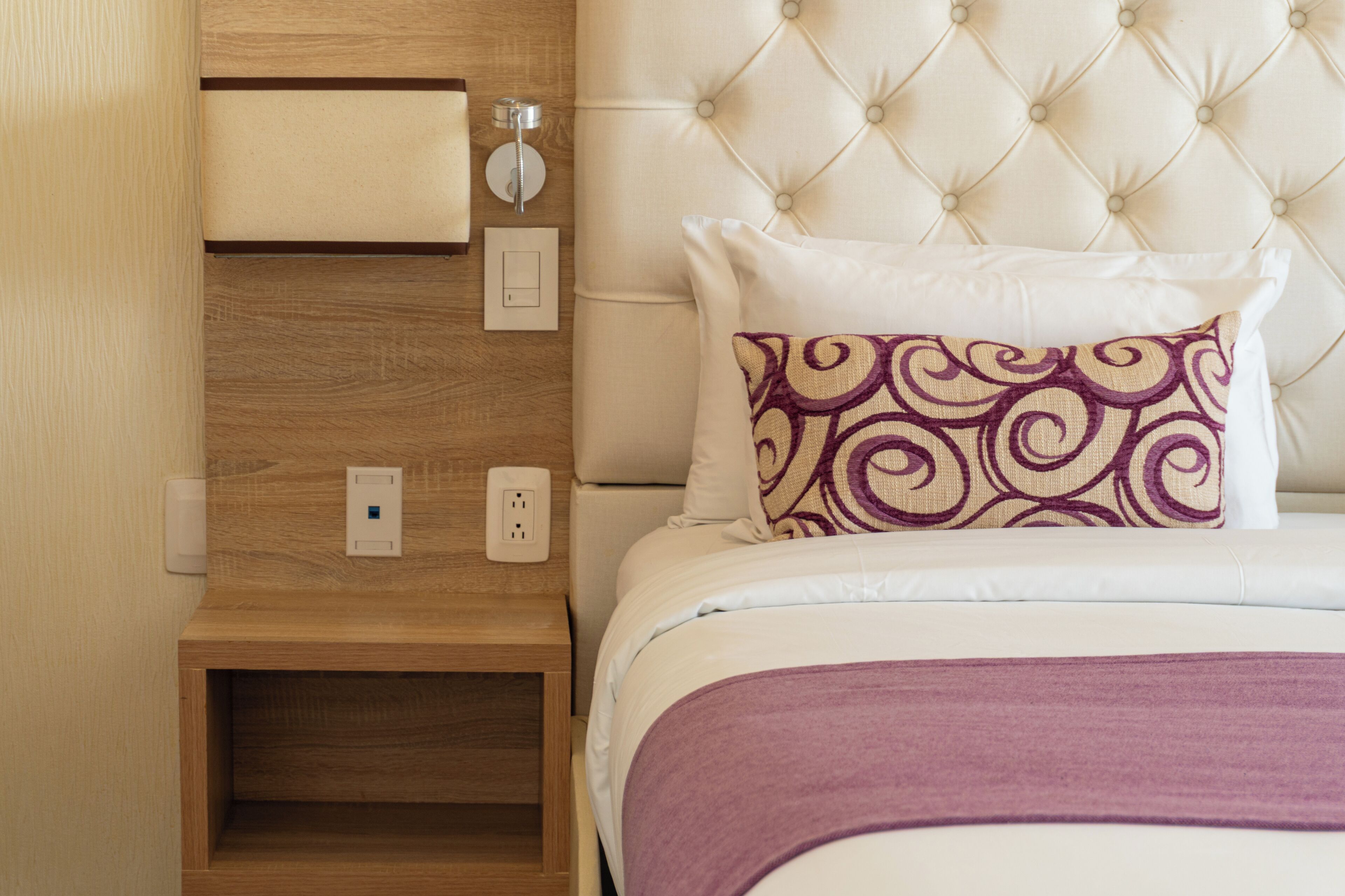 A neatly made bed with a tufted headboard, a wooden nightstand with a drawer, and a wall-mounted light switch.