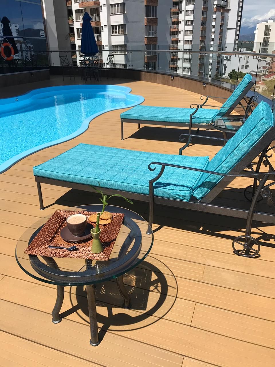 Een zwembadterras met twee ligstoelen, een tafel met een drankje en een koekje, en een woongebouw op de achtergrond.