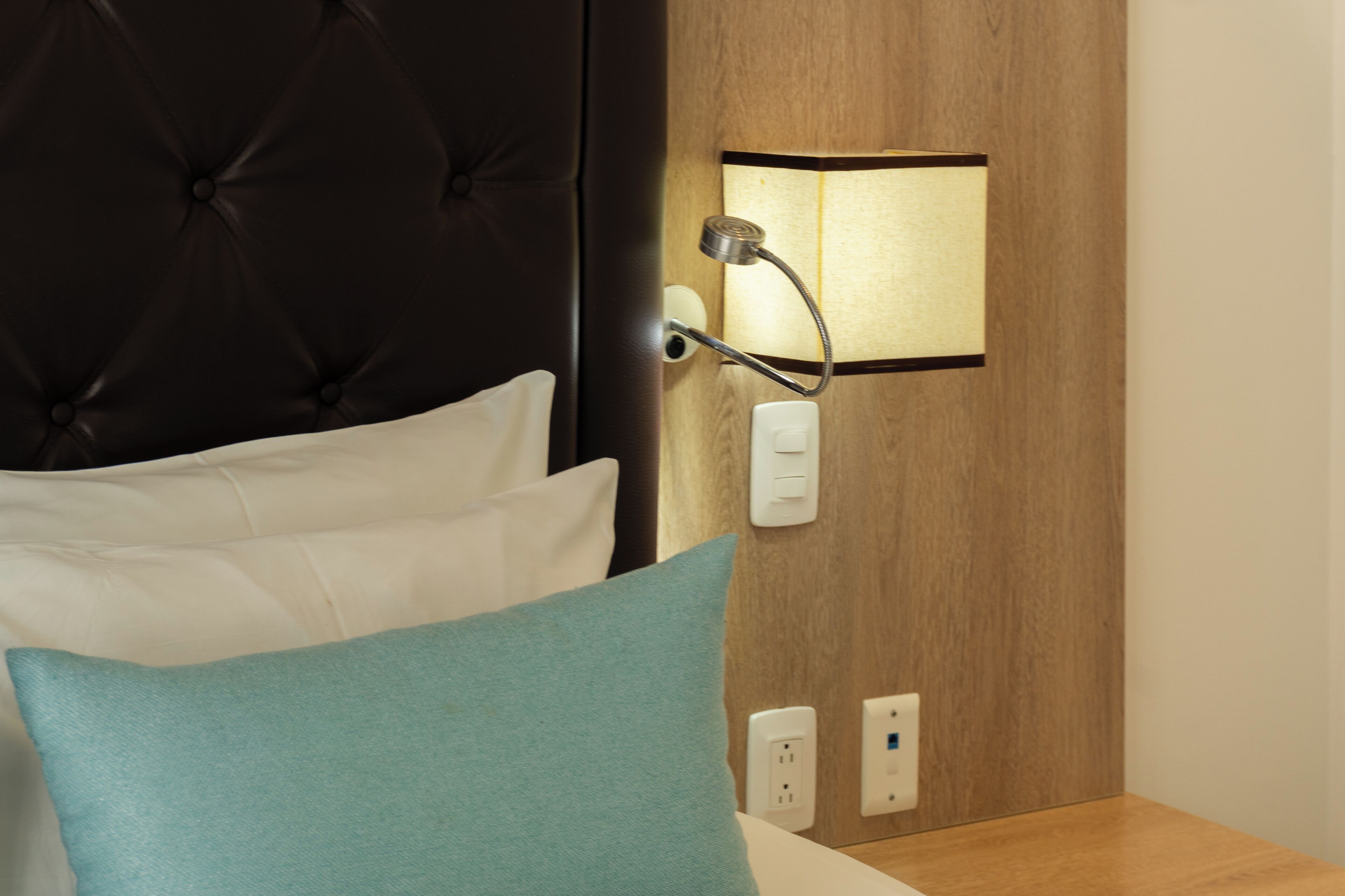 A neatly made bed with white and teal pillows, a dark headboard, and a wall-mounted light fixture with a wooden panel behind it.