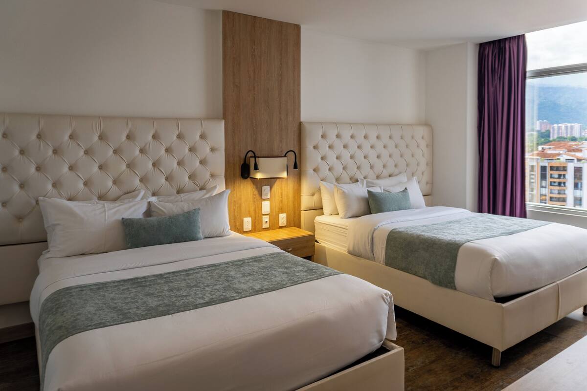 A hotel room with two beds, a wooden accent wall, a nightstand with a lamp, and a view of buildings through a window with purple curtains.