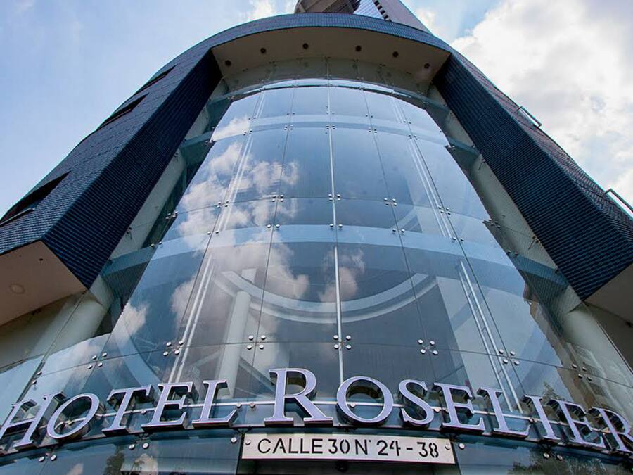 Ett modernt hotell med en böjd glasfasad, där namnet ”HOTEL ROSELIER” är tydligt synligt, och en skylt som visar adressen ”CALLE 30 N° 24-38”.