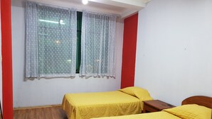 A room with two single beds, a wooden floor, a window with patterned curtains, and a red accent wall.