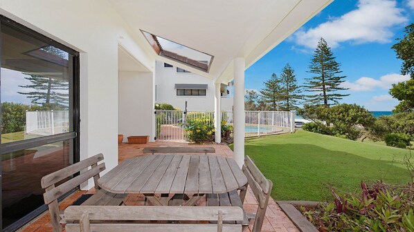 A covered patio with a wooden table and benches, overlooking a swimming pool and a well-maintained lawn with trees.