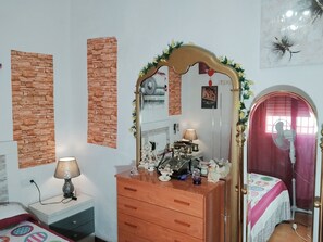 Ein Schlafzimmer mit Kommode, Spiegel, Lampe, Bett mit gemustertem Überwurf und Wand mit Ziegeloptik.