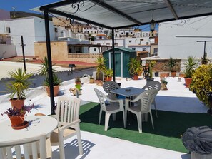 Eine Dachterrasse mit weißen Plastikstühlen und -tischen, umgeben von Blumentöpfen und einem kleinen Schuppen.