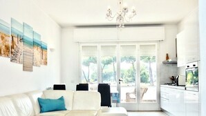 Ein modernes Wohnzimmer mit einem weißen Ecksofa, einer Deckenleuchte und einem großen Fenster mit Blick auf Bäume.