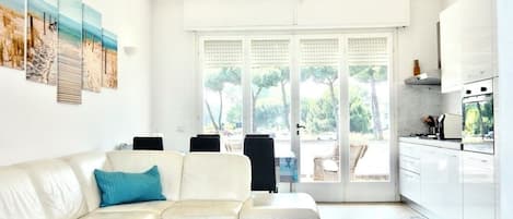 Ein modernes Wohnzimmer mit einem weißen Ecksofa, einer Deckenleuchte und einem großen Fenster mit Blick auf Bäume.