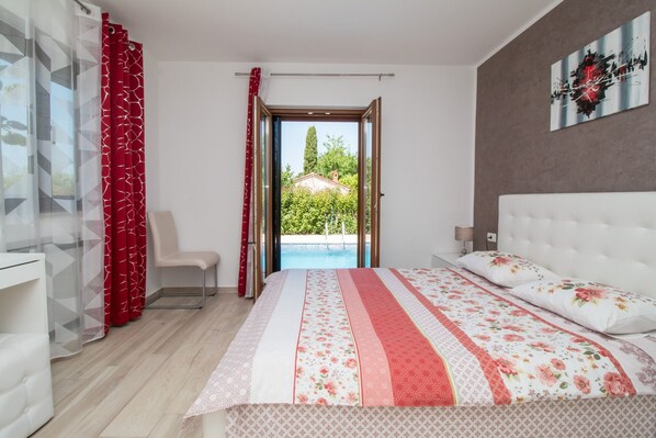 Une chambre avec un lit, une chaise, une fenêtre donnant sur une piscine et des arbres, et un tableau accroché au mur.