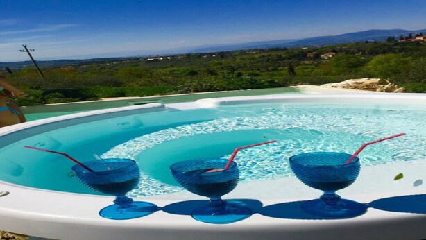 A hot tub with three glasses of blue liquid, surrounded by a pool, with a scenic view of hills and buildings in the background. - Farmhouse Il Colle (Bagno A Ripoli)