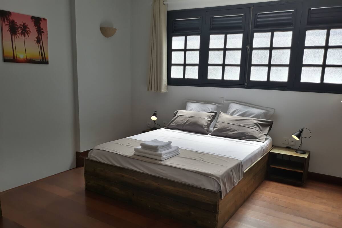 A bedroom with a wooden bed, white bedding, two bedside tables with lamps, a window with blinds, and a wall-mounted light fixture.