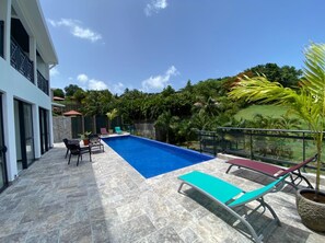 A swimming pool with lounge chairs and a table, surrounded by a stone deck and a view of greenery. - Villa Clémence (Le Francois)