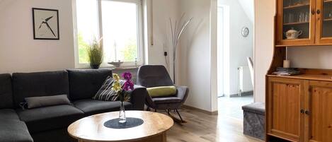 Una sala moderna con un sofá gris oscuro, una mesa redonda de madera, un sillón gris y un mueble de madera con puertas de cristal.