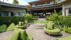 A modern outdoor seating area with green cushions, a wooden deck, and a view of a building with a covered patio. - The Elementum (Pecatu)