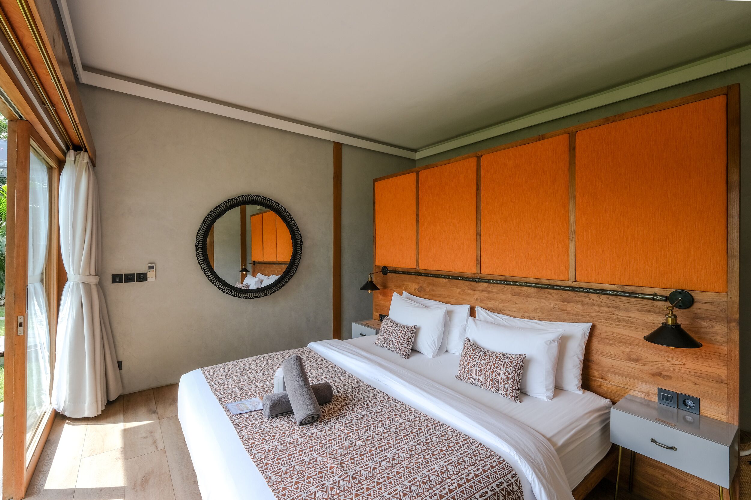 Un lit bien fait, recouvert d’une couvre-lit à motifs, une table de chevet avec une lampe et un grand miroir accroché au mur.