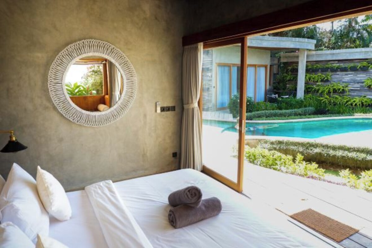 Une chambre à coucher avec un lit, des oreillers, un miroir rond et une vue sur une piscine extérieure.