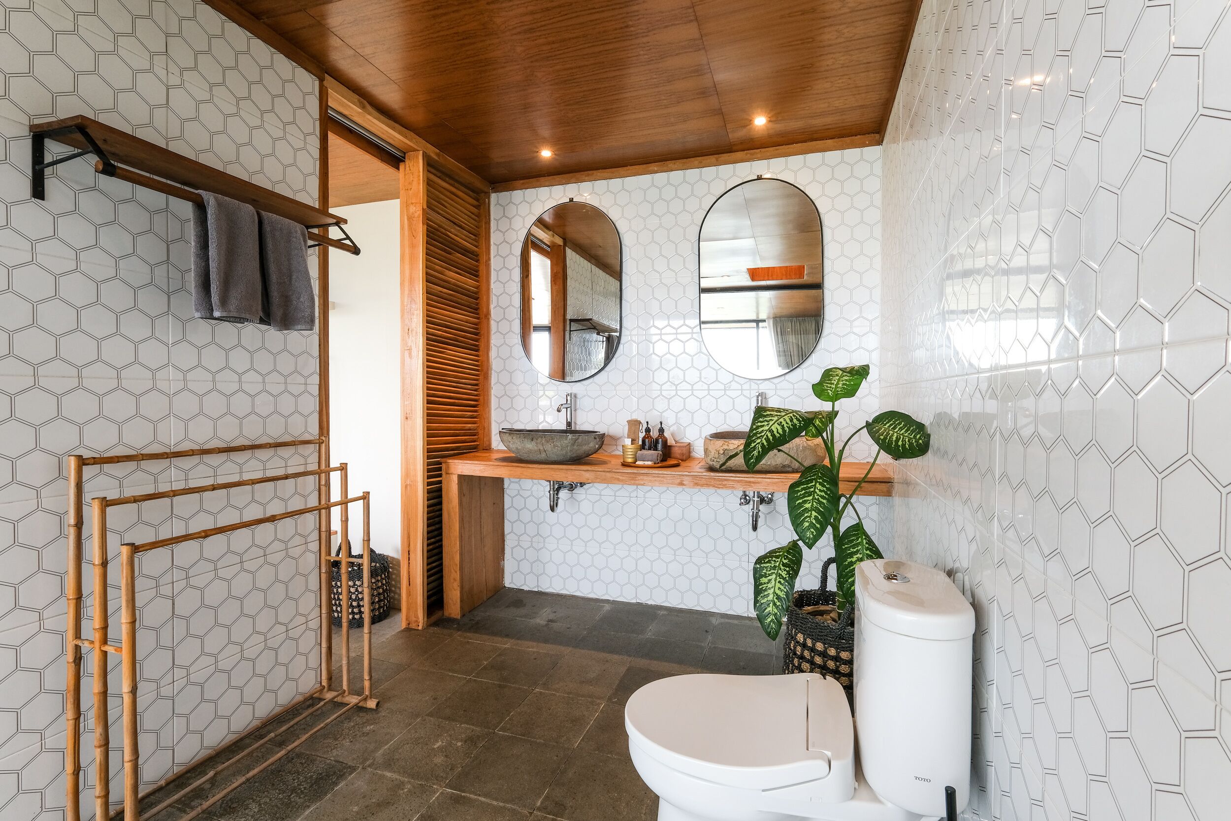 Casa de banho com painel de azulejos hexagonais, toucador de madeira, lavatório com cuba, sanita e porta-toalhas de bambu.