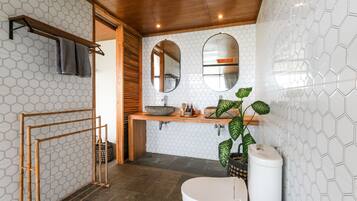 Casa de banho com painel de azulejos hexagonais, toucador de madeira, lavatório com cuba, sanita e porta-toalhas de bambu.