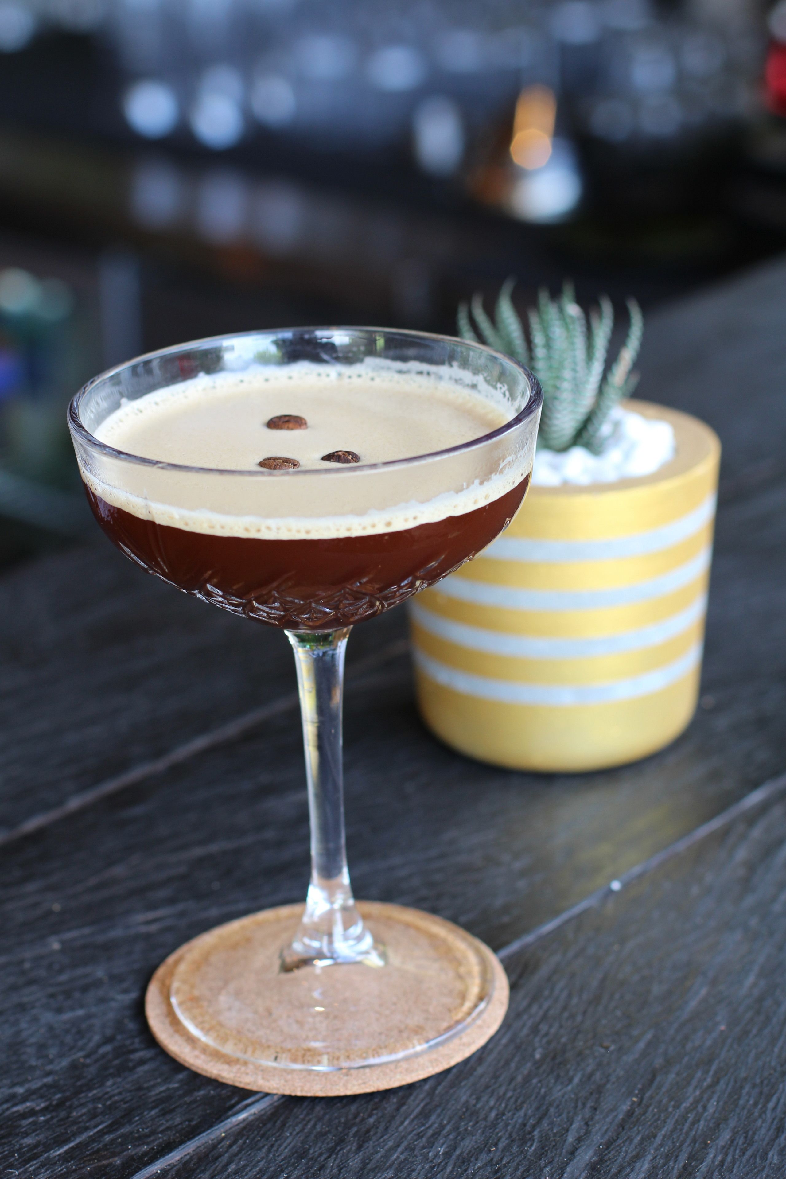 Een cocktail in een bolglas op een onderzetter, met een gestreept gele vaas en een kleine plant op de achtergrond.