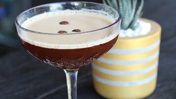 Een cocktail in een bolglas op een onderzetter, met een gestreept gele vaas en een kleine plant op de achtergrond.