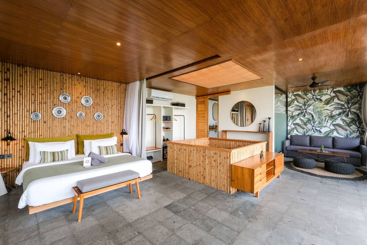 Una habitación de hotel moderna con techo de madera, una cama grande con reposabajos, un escritorio de madera, un sofá y un papel tapiz tropical.