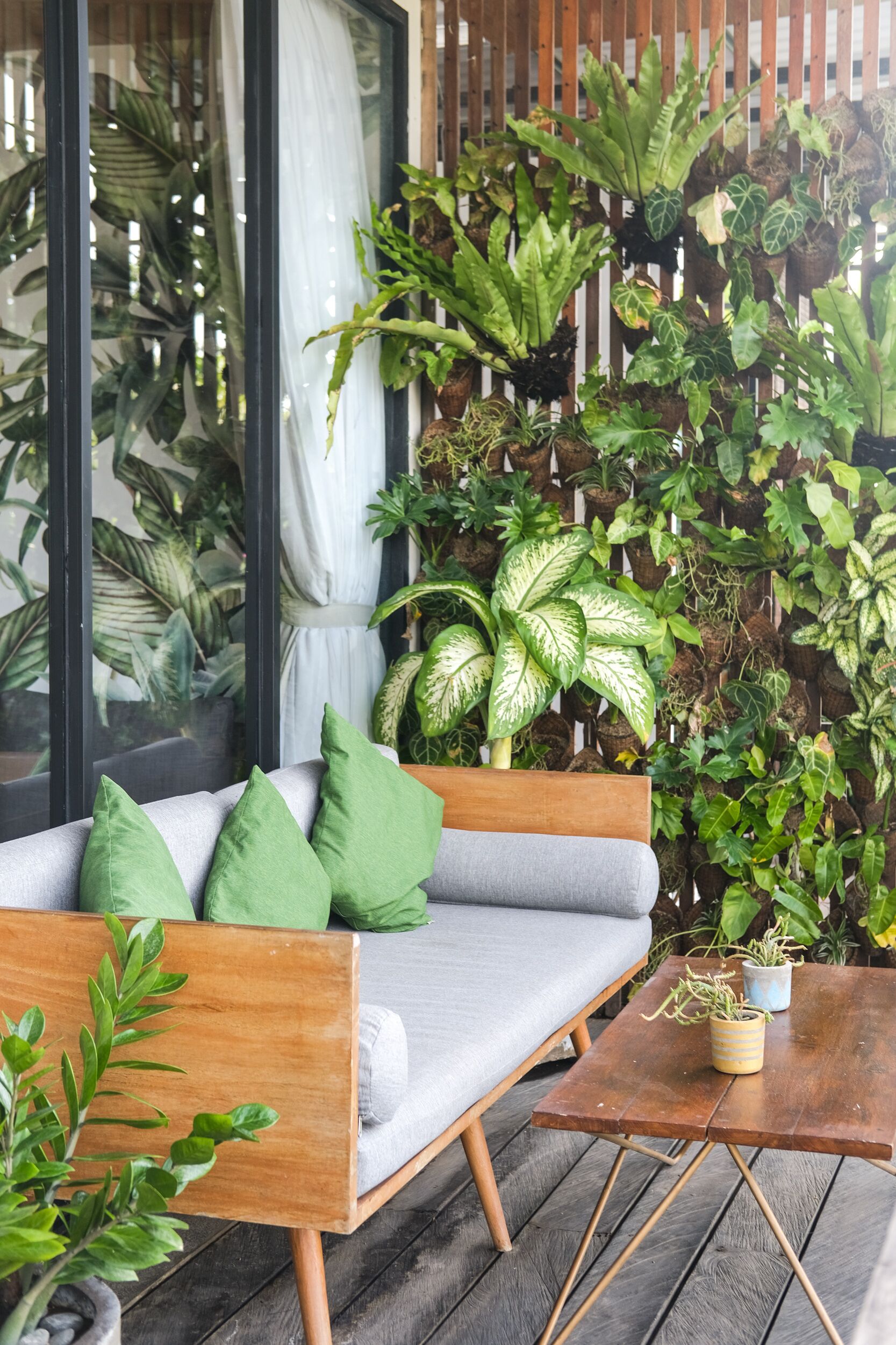 A wooden deck with a grey sofa and green cushions, surrounded by a variety of potted plants and a vertical garden.