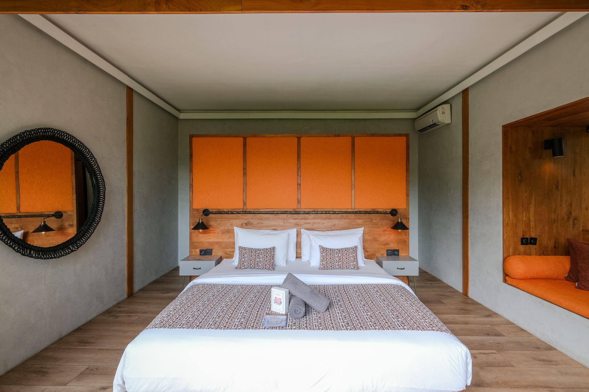 A modern hotel room with a large bed, a wooden headboard, and a small seating area with an orange cushion.