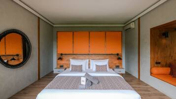 Habitación de hotel moderna con una cama grande, cabecera de madera y un pequeño rincón de descanso con un cojín naranja.