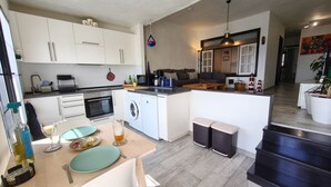 A modern kitchen with white cabinets, a built-in oven, and a washing machine. There is a dining area with a table, chairs, and a glass of wine. - Relax in front of the Ocean with a beautiful view! (Sardina del Norte)