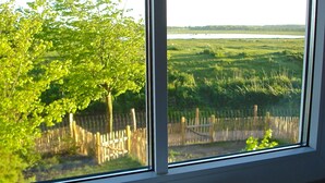 A window with a view of a green landscape, a wooden fence, and a white ceramic object on a wooden shelf.