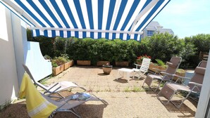 A patio with striped awning, outdoor seating, potted plants, and a building in the background. - 2 rooms cabin on the ground floor terrace (LE GRAU DU ROI)