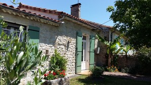Un edificio de piedra con techo de tejas, contraventanas verdes y un pequeño jardín con flores y plantas.